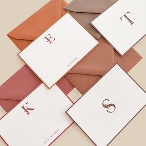 May include: Stationery set featuring personalized notecards and matching envelopes. The notecards have a white background with a single initial and name printed in a dark reddish-brown color. The envelopes are in shades of brown and red.