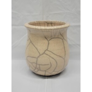 May include: A cream-colored ceramic vessel with a wide, rounded body and a flared rim. The exterior surface features a distinct dark gray crackle glaze pattern that creates a network of lines across the entire outer body of the pottery piece.