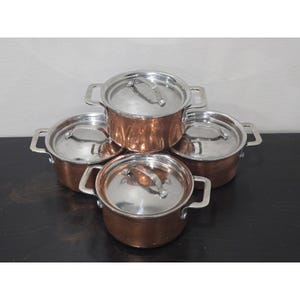 May include: A set of four round copper pots with silver-toned metal lids and side handles. Each pot features a polished metallic finish on the lid and a warm copper exterior. The pots are arranged in a cluster, showcasing their matching design.