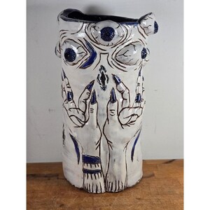 May include: A cylindrical ceramic vase with a white glaze and dark blue accents. The surface features sculpted relief details including multiple eyes near the rim and two hands with long, pointed fingers reaching upward toward a central eye motif.