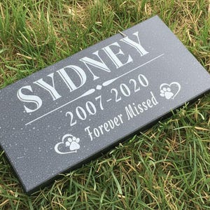 May include: A dark grey rectangular pet memorial stone, inscribed with the name "SYDNEY" in white, the years "2007-2020", and the words "Forever Missed". Features paw prints and heart symbols.