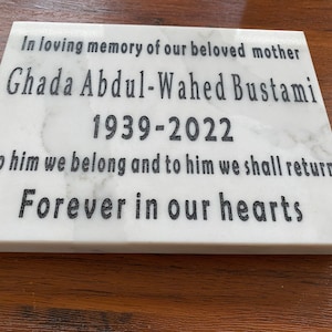 Puede incluir: Placa conmemorativa rectangular de mármol blanco con texto negro grabado. El texto dice: "En memoria cariñosa de nuestra amada madre Ghada Abdul-Wahed Bustami 1939-2022. A él pertenecemos y a él volveremos Por siempre en nuestros corazones."