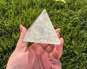 May include: A clear, crystalline pyramid is held in a hand against a backdrop of green grass. The pyramid has a multifaceted surface, catching the light. The pyramid is a geometric shape, and the overall aesthetic is natural and minimalist.