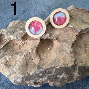 May include: Two round wooden earrings with a red and white resin inlay. The earrings are on a brown rock surface.
