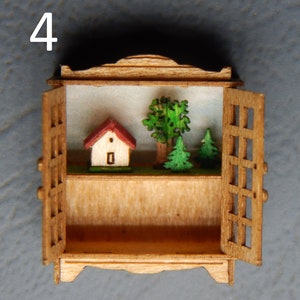 May include: A miniature wooden cabinet with a hinged door, open to reveal a small house and three trees inside. The cabinet is painted in a light wood tone and has a decorative top.