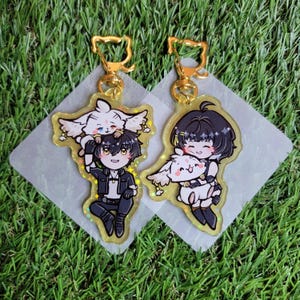 May include: Two anime-style acrylic keychains with gold-coloured cat-shaped clasps. Each keychain features a cartoon character with a small white animal. The keychains are on a green grassy surface.