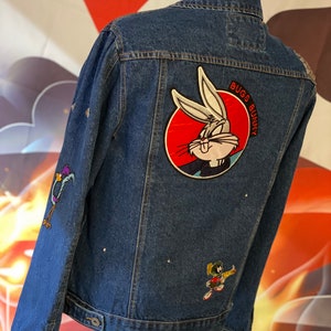 May include: A dark blue denim jacket featuring a large circular embroidered patch of Bugs Bunny on the back, with the text BUGS BUNNY. The jacket also includes smaller embroidered patches of the Road Runner on the sleeve and Marvin the Martian.