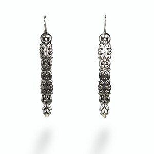 May include: A pair of long, silver-colored dangle earrings. Each earring features an ornate, vertical design with repeating floral and scrollwork patterns. The earrings have a hook closure and are displayed against a white background.