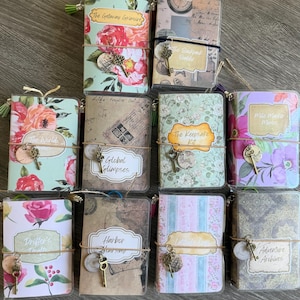 May include: A collection of small, decorative journals with floral and patterned covers. Each journal has a unique title, such as "The Getaway Grimoire" and "Adventure Archives", and is adorned with a key charm and tassel.