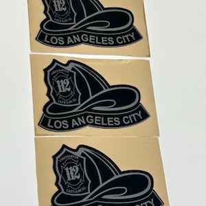 May include: Three rectangular stickers featuring a black graphic of a firefighter helmet with the number 112, the text FIREFIGHTER IAFF-LOCAL, and a banner at the bottom reading LOS ANGELES CITY, all set against a light tan adhesive backing.