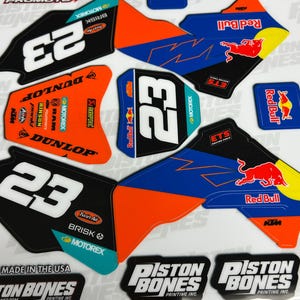 May include: A set of motocross decals featuring the number 23, the words "Red Bull", "Piston Bones", and "Dunlop", and various sponsors' logos. The decals are printed on a white background and are designed to be applied to a motorcycle.