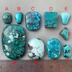 May include: A selection of turquoise gemstones in diverse forms, such as oval, rectangular, and triangular. The stones exhibit a spectrum of blue-green shades with dark veining. A ruler with centimetre measurements is present.