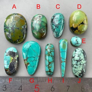 Puede incluir: Una colección de diez cabujones de turquesa pulidos etiquetados de la A a la J, con varios tonos de verde y azul con patrones de matriz oscuros. Una regla de metal está colocada en la parte inferior, mostrando medidas en milímetros.