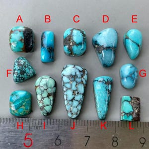May include: A collection of twelve polished turquoise cabochons in various shapes and shades of blue and green with dark matrix patterns, labelled A through L, positioned above a metal ruler showing measurements in centimetres and millimetres.