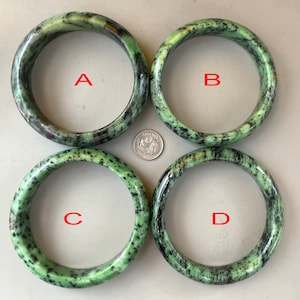 May include: Four green and black stone bangles are shown, each with a unique pattern. The bangles are circular and appear to be made of polished stone. A coin is placed in the center for size comparison. The bangles are labelled A, B, C, and D.