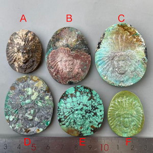 May include: Six oval-shaped, carved stone pendants with a face design. The stones vary in color, including brown, turquoise, and green. A ruler is placed below the pendants, with measurements in inches.