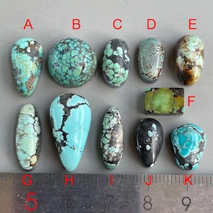 Può includere: Una collezione di undici cabochon di turchese lucidati etichettati da A a K, con varie tonalità di blu, verde e motivi a matrice marrone. Un righello di metallo posizionato in basso mostra che le pietre misurano da 1,5 a 4 cm di dimensione.