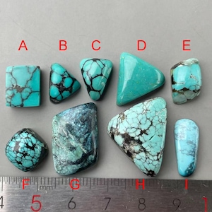 May include: A collection of turquoise gemstones in diverse shapes, such as rectangular, triangular, and rounded. The stones exhibit a vivid turquoise hue with dark matrix patterns. A ruler with millimeter markings is positioned beneath the stones.