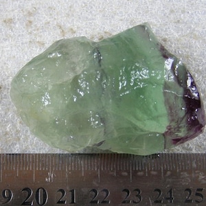 Könnte beinhalten: Ein roher Fluoritkristall mit Grün- und Lilatönen. Der durchscheinende Stein hat eine kristalline Struktur und liegt neben einem Metalllineal. Fluorit ist ein natürliches Mineral, das oft in Schmuck und Dekoration verwendet wird.