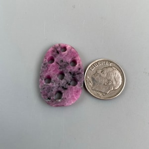 May include: A pink and black oval-shaped stone with holes drilled through it. The stone is about the size of a US dime.