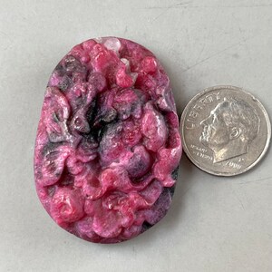 Puede incluir: Un colgante tallado de piedra preciosa rosa y gris con un diseño floral. El colgante es aproximadamente del tamaño de una moneda de diez céntimos de euro.