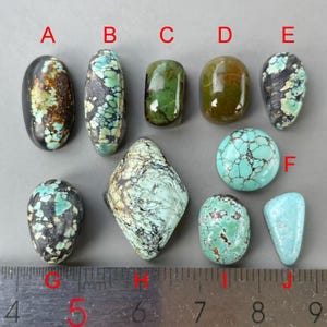 Puede incluir: Una colección de diez cabujones de turquesa pulidos etiquetados de la A a la J, dispuestos sobre una regla de metal. Las piedras varían en forma, incluyendo ovalada, redonda y de diamante, con diversos patrones de vetas de matriz azules, verdes, marrones y negras.