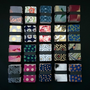 May include: A collection of small, rectangular notebooks arranged in a grid. Each notebook is secured with a black band and has a distinct patterned cover. The covers showcase diverse designs, including floral, geometric, and abstract patterns in various colours.
