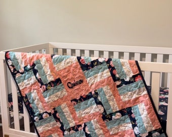May include: A pink, white, and blue floral baby quilt with a navy blue border. The quilt is personalized with the name "Quinn" embroidered in the center. The quilt is draped over a white crib.