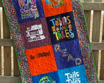 May include: A colorful quilt made from repurposed t-shirts featuring a variety of reading-themed designs. The quilt is bordered with a floral print fabric and has a green trim. The quilt squares feature the text "The more you read the more you learn", "Library Rock", "No Shelf Control", "Tails and Tales", "Read", and "Reading Rocks!"