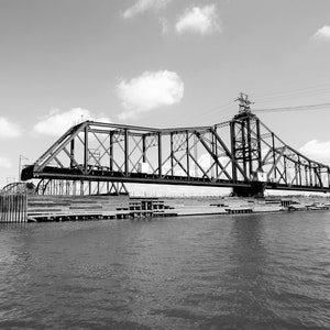 May include: A black and white photo of a large, metal drawbridge over a body of water. The bridge is raised, allowing boats to pass underneath. The bridge is made of steel beams and has a series of vertical supports.