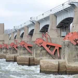 May include: A concrete dam with multiple red metal gates. The gates are raised and the water is flowing over the dam.