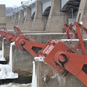 May include: A close-up of a dam with several red metal gates. The gates are partially open and water is flowing through them. The dam is made of concrete and has a bridge over it. The water is frozen in places.