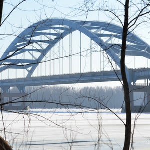 May include: A view of a large, steel arch bridge over a frozen river. The bridge is gray and has a series of vertical supports. The river is covered in snow and ice. Bare trees are in the foreground.