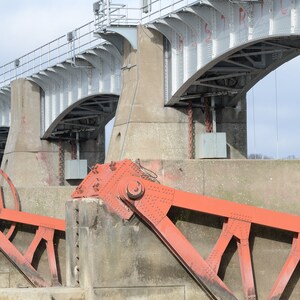 May include: A close-up of a large, gray concrete dam with red metal gates. The gates are partially open, revealing the rushing water below. The dam is made of concrete and steel, and it has a series of arches that support the structure.