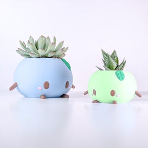 May include: Two whimsical, round planters in light blue and green, each with a succulent plant. The planters have brown arms and legs, pink cheeks, and a green leaf accent. The blue planter holds a large succulent, while the green one has a smaller, spiky plant.