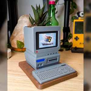 May include: A gray 3D-printed planter shaped like a vintage computer monitor with a succulent inside. The screen displays the Microsoft Windows 98 logo. A matching gray keyboard sits in front of the monitor base on a brown wooden rectangular platform.