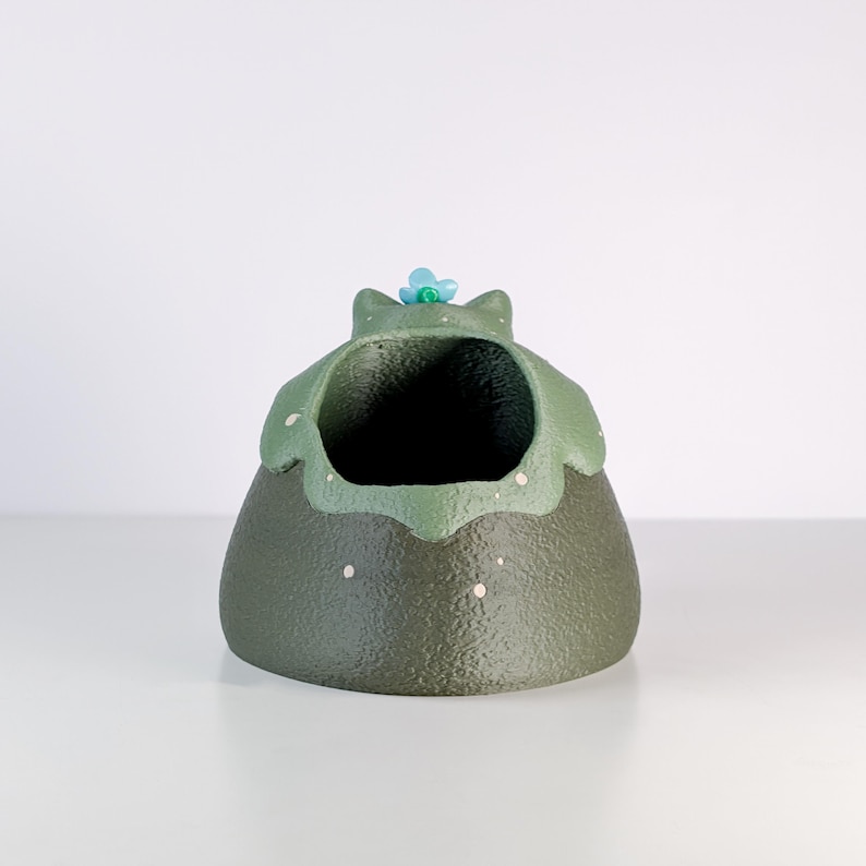 May include: A ceramic planter in the shape of a frog. The planter is primarily olive green with a lighter green rim and a small blue flower on top. White speckles dot the surface.
