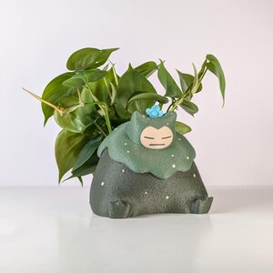 May include: A dark green ceramic planter shaped like a cartoon character, with a light green collar dotted with white spots. The planter holds a green leafy plant. A small blue flower decorates the top of the planter.
