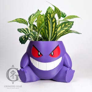 May include: A purple planter shaped like a cartoon character with red eyes and a wide grin, holding green and yellow-striped plants. The planter has a rounded body and small feet. The logo "Grimoire Gear" is visible in the lower left corner.