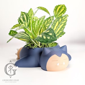 May include: A navy blue plant pot in the shape of a sleeping cartoon character, with a light tan face and white teeth. The pot contains a green plant with yellow-green leaves and moss. The logo "Grimoire Gear" is at the bottom.