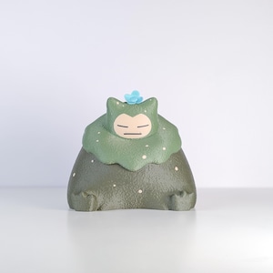 May include: A green and gray figurine of a character with closed eyes and a small blue flower on its head. The figurine has a round body and a head that resembles a cat. The character is wearing a green leafy garment.