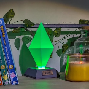 May include: A green diamond-shaped lamp with a grey base and the name "Olivia" on a small plaque. The lamp is lit, casting a green glow. A lit candle and books are in the background.