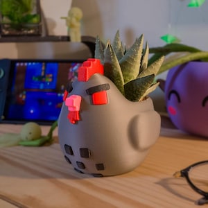 May include: A gray, chicken-shaped planter with a succulent plant inside. The planter features a red comb, a pink beak, red eyes with black eyebrows, and black square markings on the body. The design has a matte finish and rounded features.