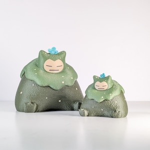 May include: Two green and white figurines, resembling a character with a round body and a leafy head. The larger figure is in the foreground, with a smaller version behind it. Each has a blue flower on its head.
