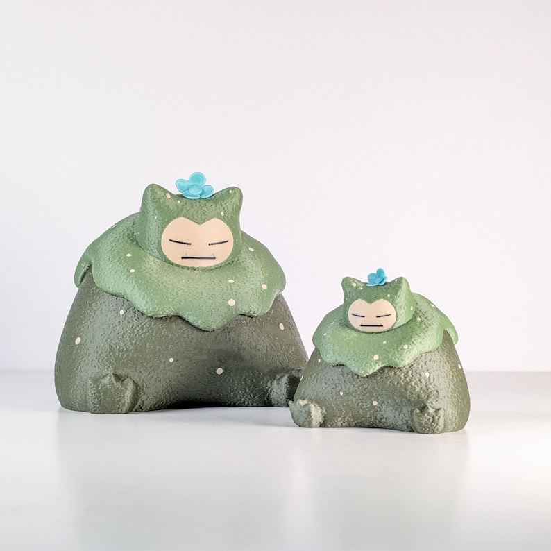May include: Two green and white figurines, resembling a character with a round body and a leafy head. The larger figure is in the foreground, with a smaller version behind it. Each has a blue flower on its head.