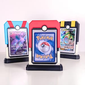 May include: Three Pokémon card display stands in blue, red, and yellow. Each stand has a black base, a white frame, and a coloured top with a white circular accent. The stands hold Pokémon trading cards, displaying the card's front and back.