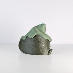 May include: A ceramic frog-shaped container in shades of green and dark olive. The frog has a textured surface with white speckles and a light blue accent. The container is sitting on a white surface.