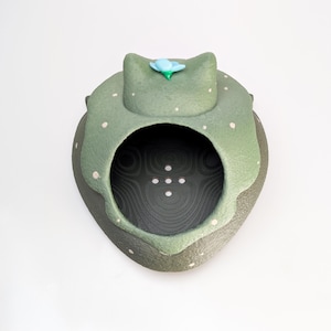 May include: A handcrafted ceramic sculpture in the shape of a frog. The frog is primarily green with white polka dots and a blue flower on its head. The interior is dark with a textured pattern.