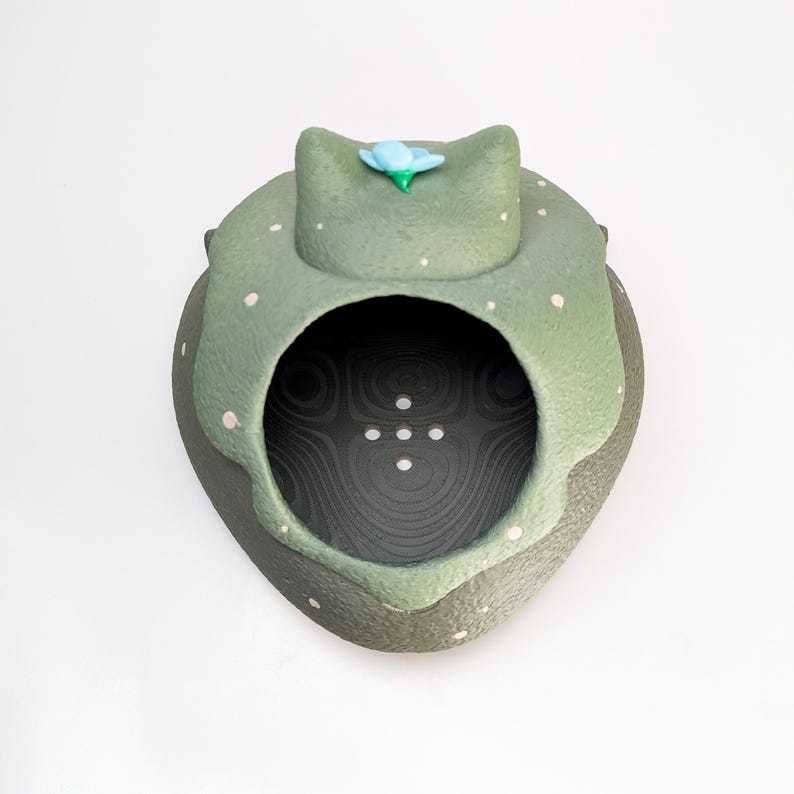 May include: A handcrafted ceramic sculpture in the shape of a frog. The frog is primarily green with white polka dots and a blue flower on its head. The interior is dark with a textured pattern.