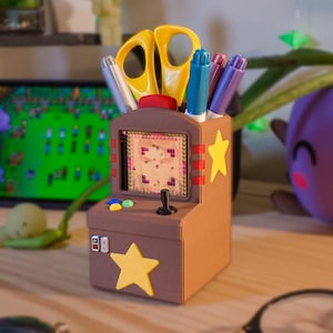 May include: A brown desk organizer shaped like a miniature arcade cabinet with a screen displaying a game, a black joystick, and colorful buttons. The unit features yellow star accents and holds yellow scissors, a blue marker, and two purple markers.